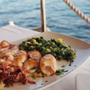 Grilled squid with boiled potatoes and chard (blitva) on white plate with sea on background. Traditional Croatian food. Rovinj, July 2017.